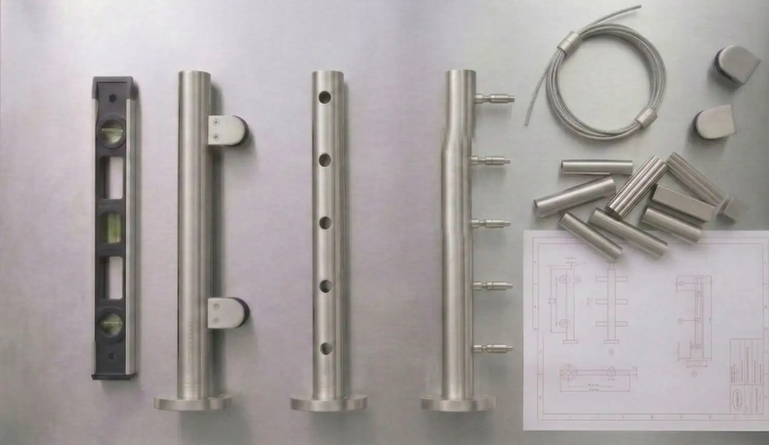 Flat-lay of three stainless steel stanchion variants — glass clip stanchion, round tube stanchion, and cable stanchion — with spirit level, cable fittings, and installation plans on a brushed steel surface